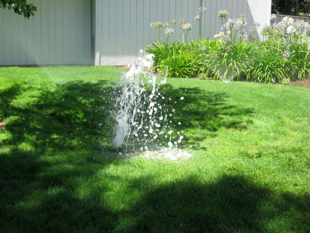 Picture of a green lawn with 2 functioning sprinklers and 1 broken sprinkler that is wasting water.