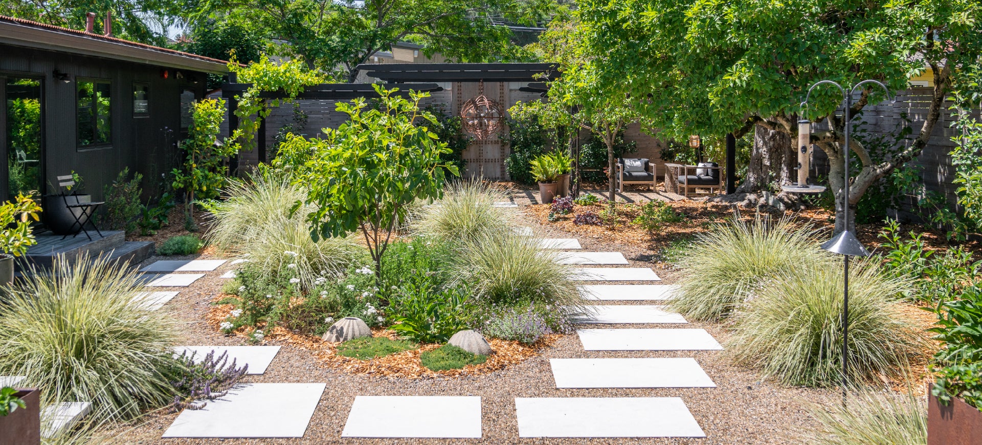 low-water use landscaped backyard with modern concrete step pads set in pea gravel, a pergola with seating in the background, house painted black on the left, wood fence on the right