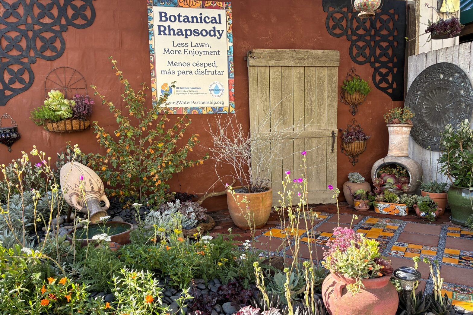Waterwise demonstration garden with colorful tile patio, California native plants and many potted succulents. Sign says 'Botanical Rhapsody. Less Lawn more enjoyment. Menos cesped, mas para disfrutar'