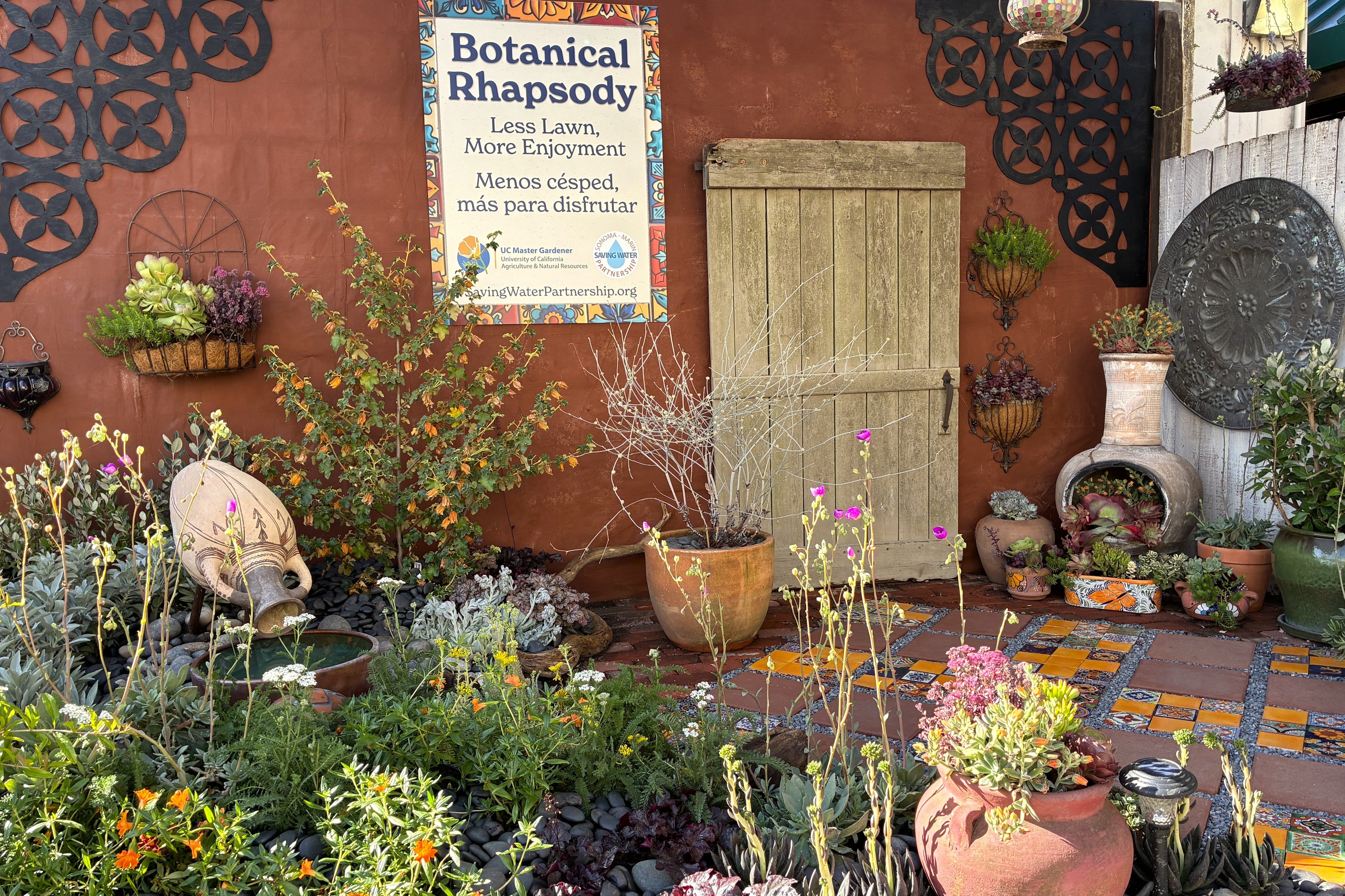 Waterwise demonstration garden with colorful tile patio, California native plants and many potted succulents. Sign says 'Botanical Rhapsody. Less Lawn more enjoyment. Menos cesped, mas para disfrutar'
